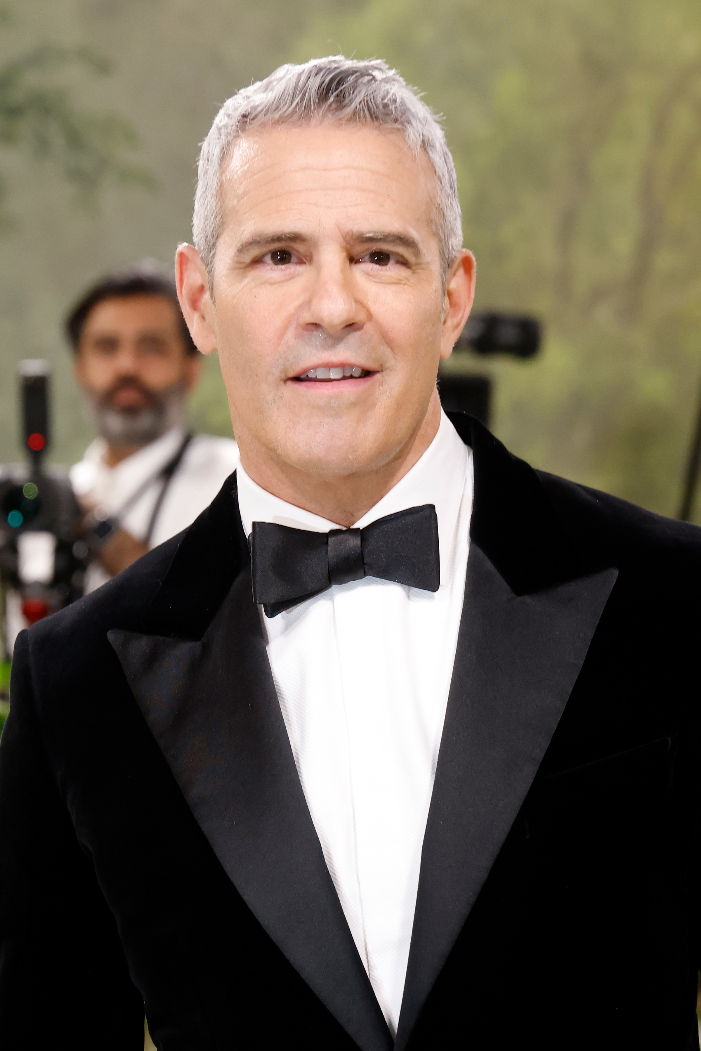 Andy Cohen wearing a tuxedo with a bow tie, smiling at the camera with a blurred background and a photographer behind him