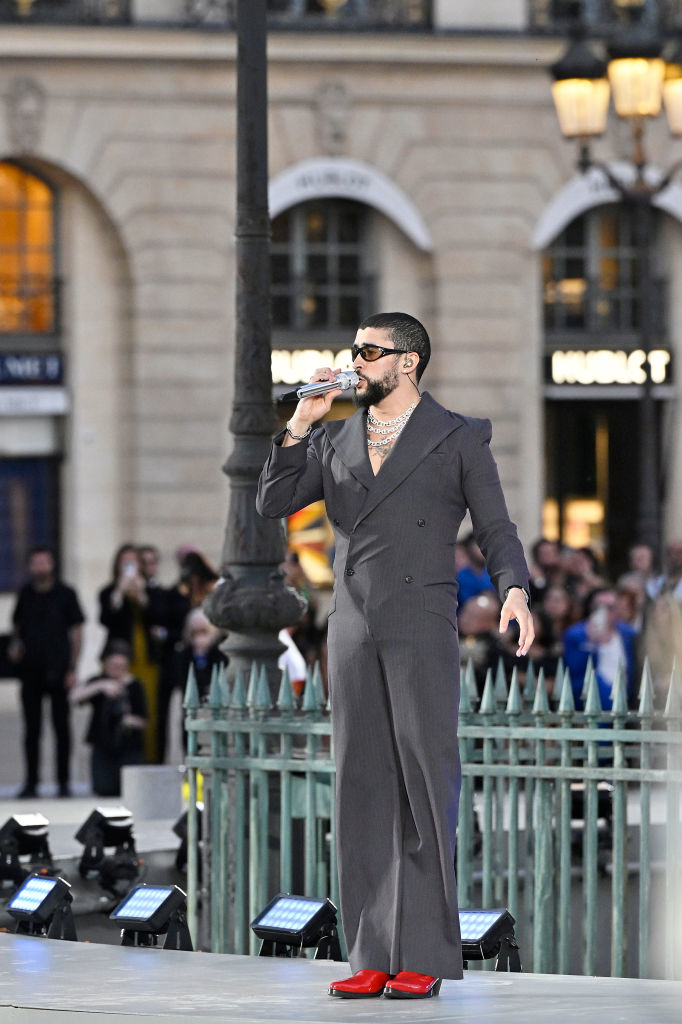 Bad Bunny, in a stylish dark suit and red shoes, performs on stage at an outdoor event, holding a microphone