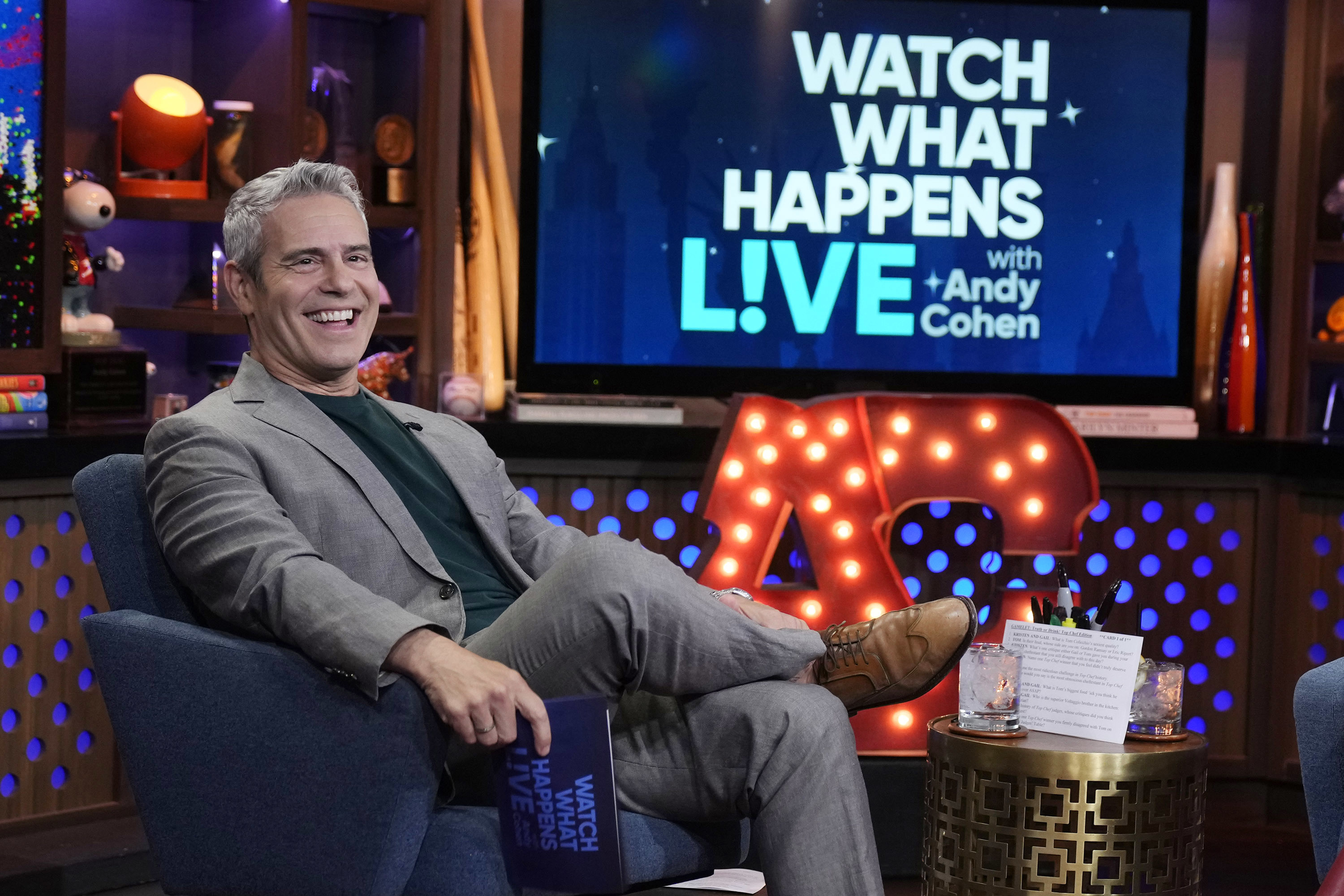 Andy Cohen sits smiling on a TV set holding cue cards for "Watch What Happens Live with Andy Cohen." The set is stylish with a large illuminated "AC" sign