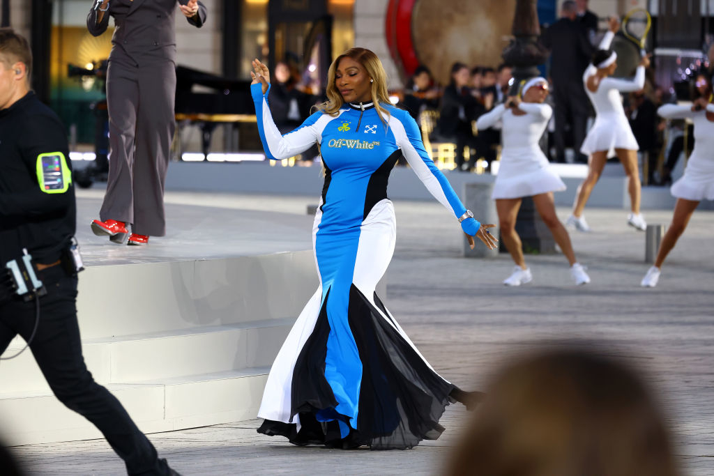 Serena Williams is walking in a long, form-fitting dress with multiple panels on a stage with performers in the background