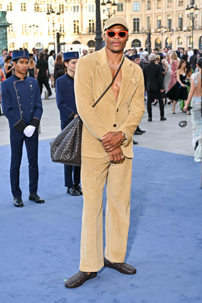 Russell Westbrook in a tan corduroy suit, sunglasses, and carrying a black bag at an event with uniformed staff and attendees in the background