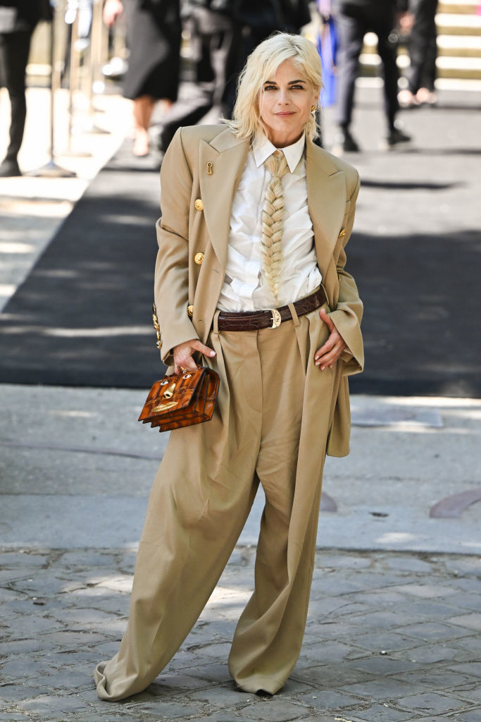 Selma Blair wears an oversized tan suit, white shirt with a twisted front, and holds a small handbag at a public event