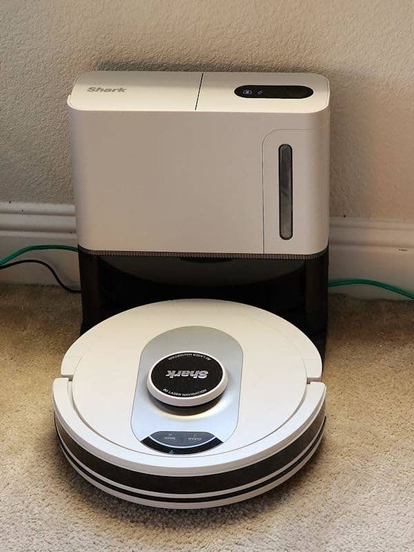 A Shark robotic vacuum cleaner with its charging base is placed on a carpeted floor near a wall