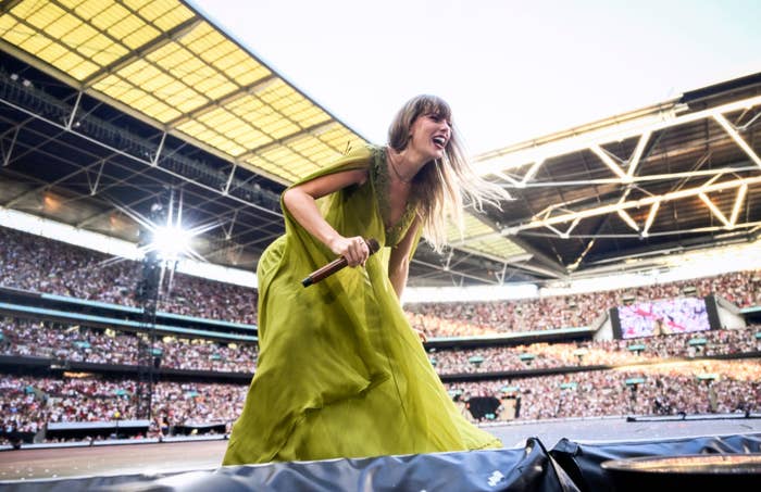 Taylor Swift performing on stage in a flowing dress at a large stadium filled with fans. She is holding a microphone and smiling