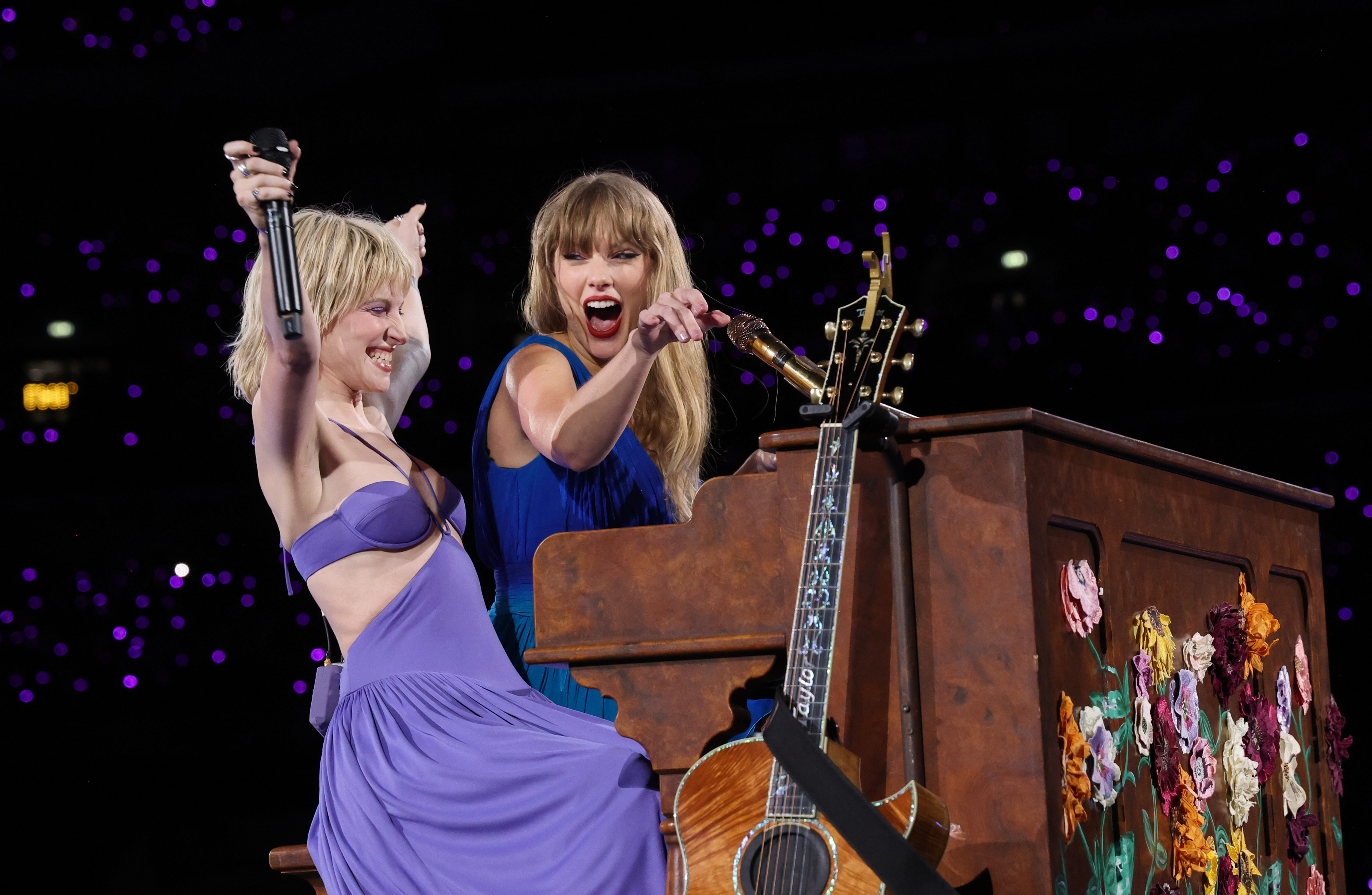 Taylor Swift and Hayley Williams on stage, Hayley wearing a purple dress while Taylor plays a flower-adorned piano and holds a guitar during a performance