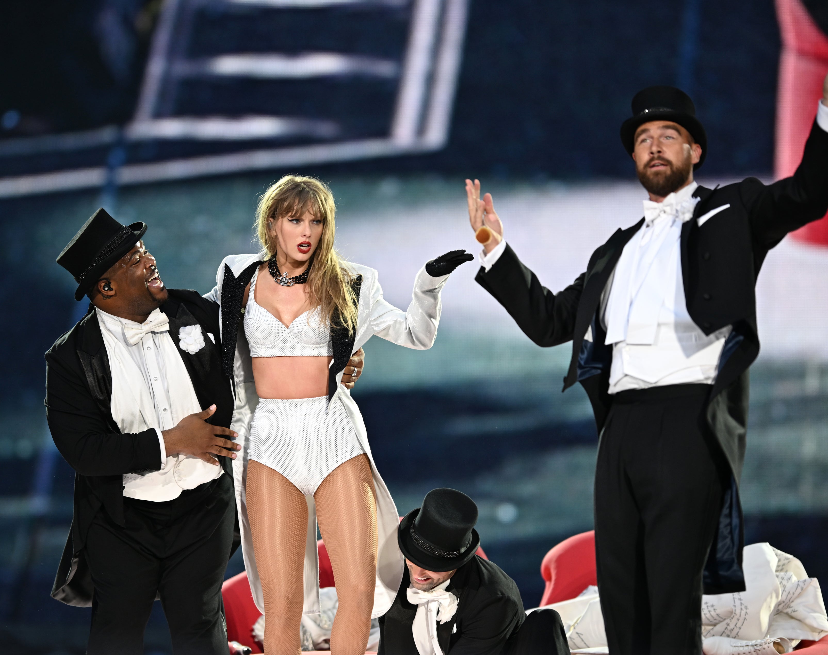 Taylor Swift in a white outfit with three dancers in tuxedos performing on stage alongside Travis Kelce