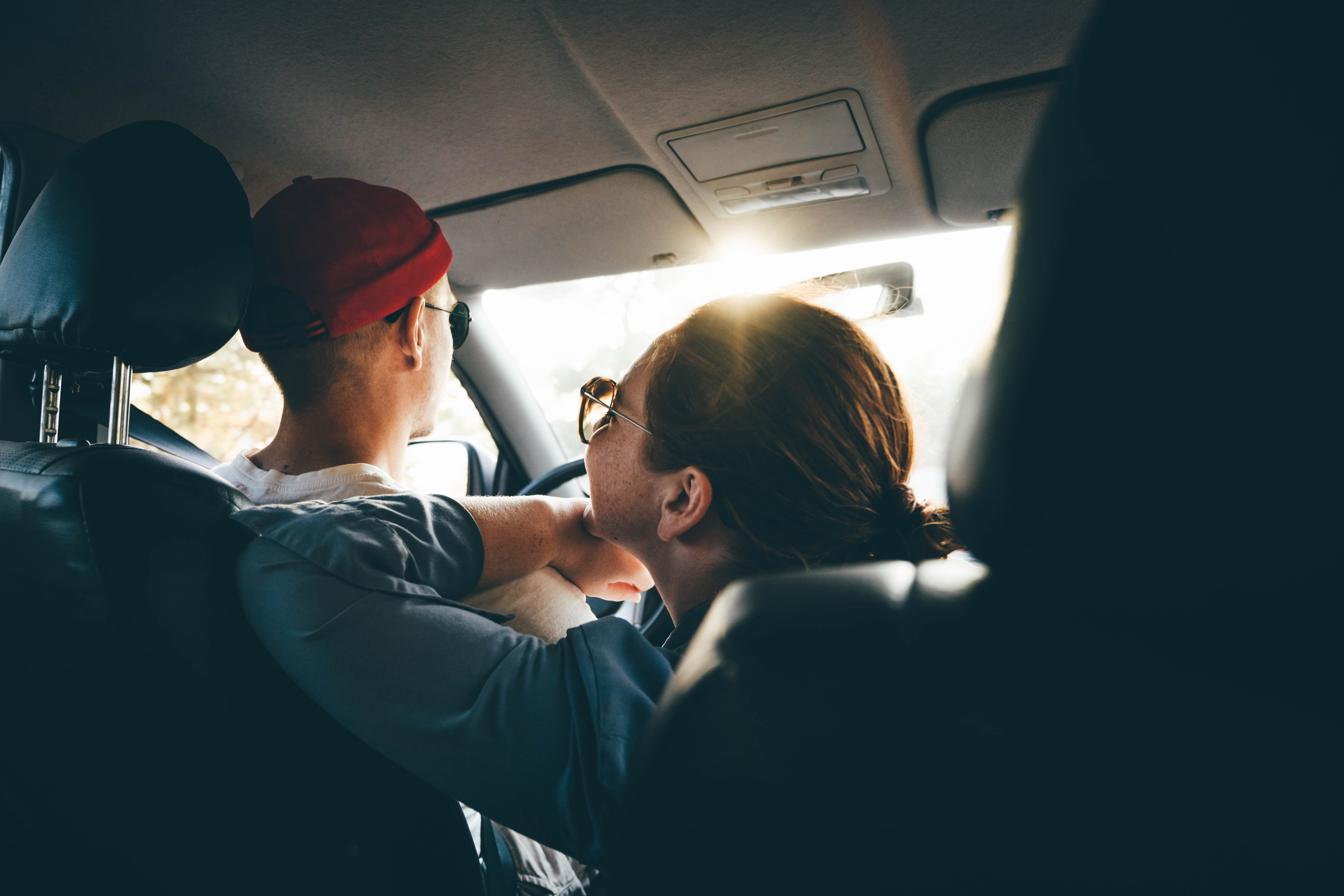 A couple, one wearing glasses and a red cap, the other leaning over the car&#x27;s console, share a close moment inside a car