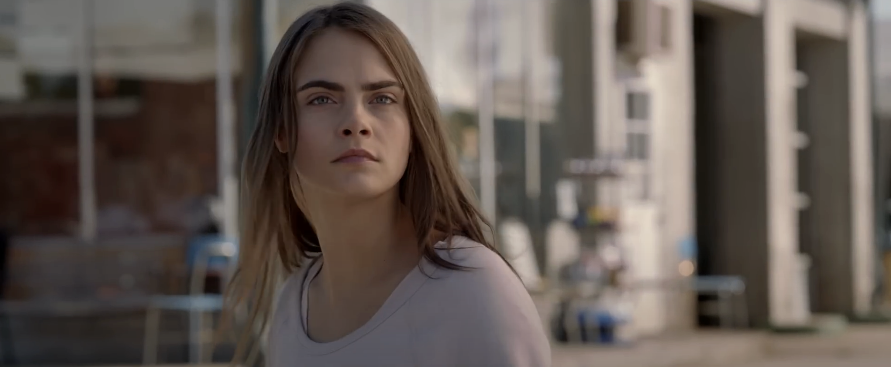 Cara Delevingne looks off-camera in a scene from the film "Paper Towns." She wears a casual, light-colored top and has long hair gently draped over her shoulders