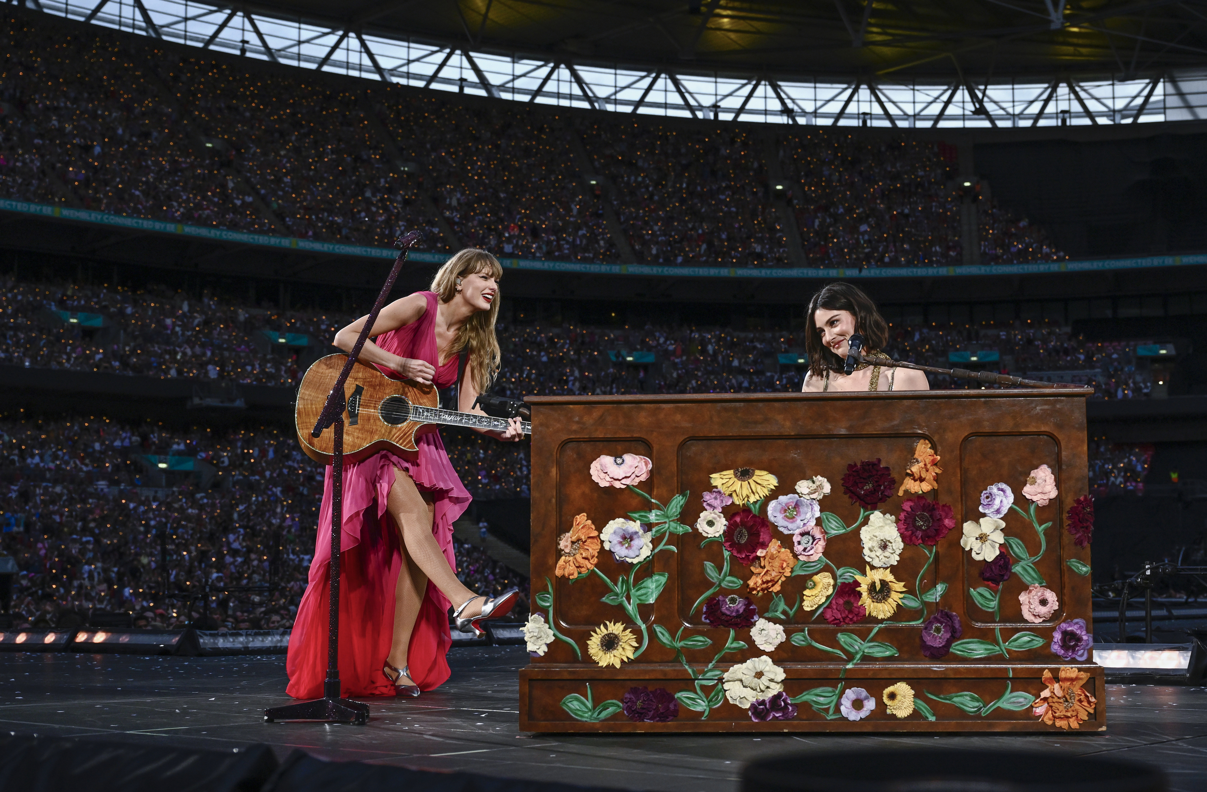 Taylor Swift in a red dress playing guitar and joined by Grace Abrams at a flower-decorated piano on stage during a concert at a large stadium