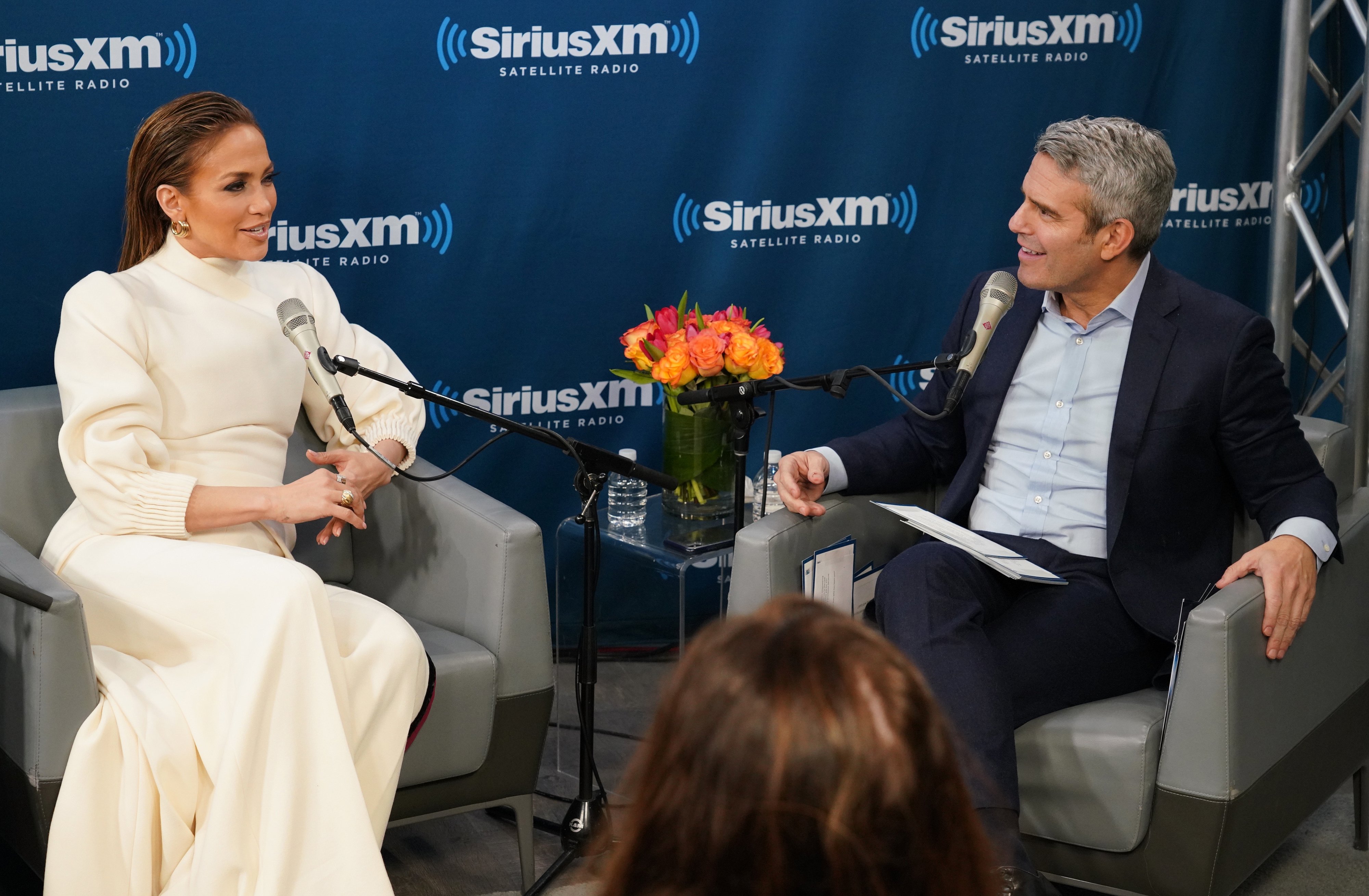 Jennifer Lopez in a chic, long-sleeved white dress being interviewed by Andy Cohen at a SiriusXM event