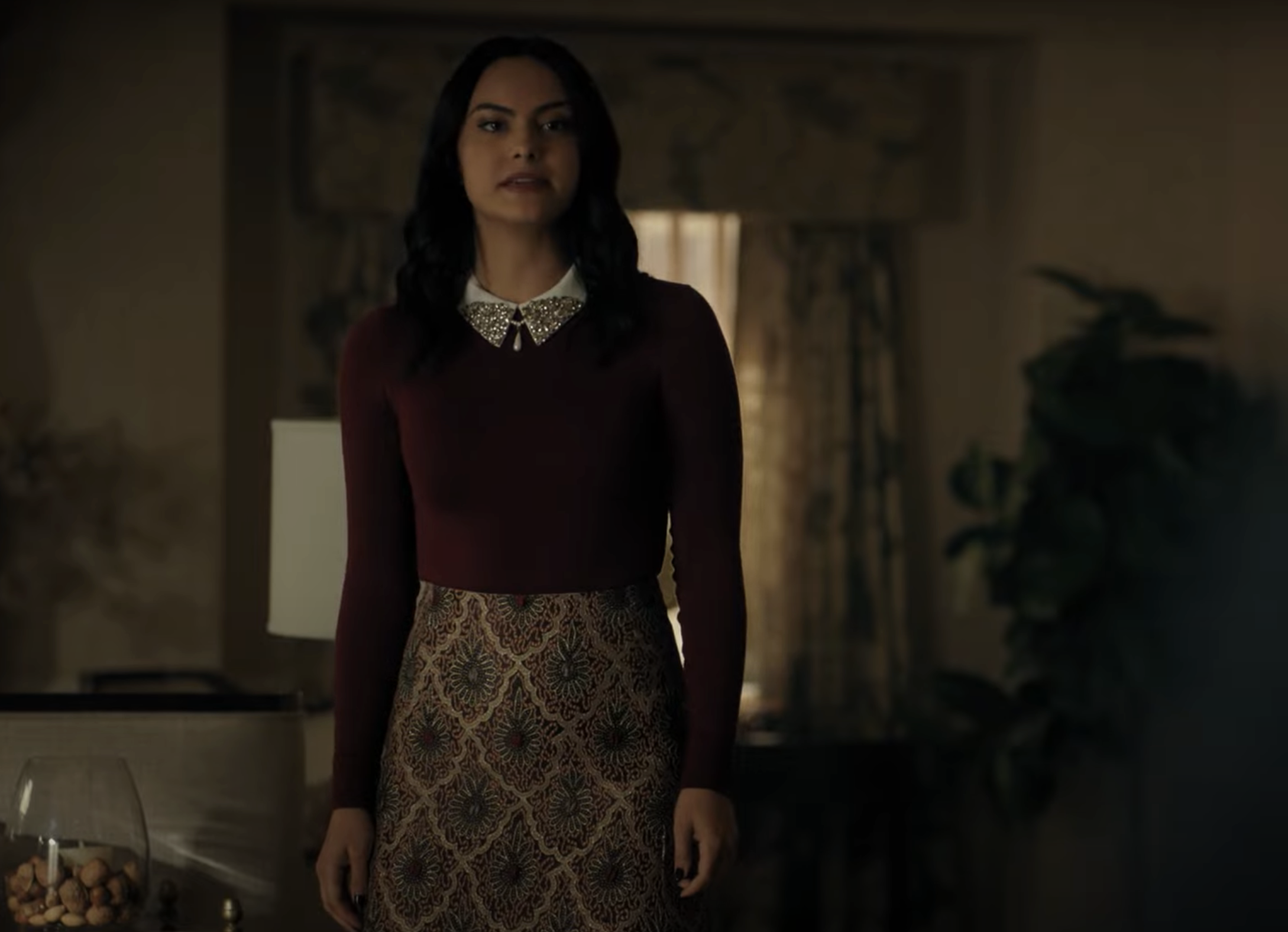 Camila Mendes stands indoors wearing a long-sleeved top with a collar and a patterned skirt