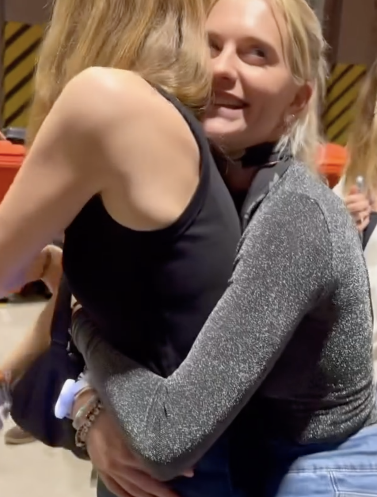 Poppy Delevingne hugging Leslie Mann, one in a sleeveless top and the other in a glittery long-sleeve shirt. Their faces are not fully visible