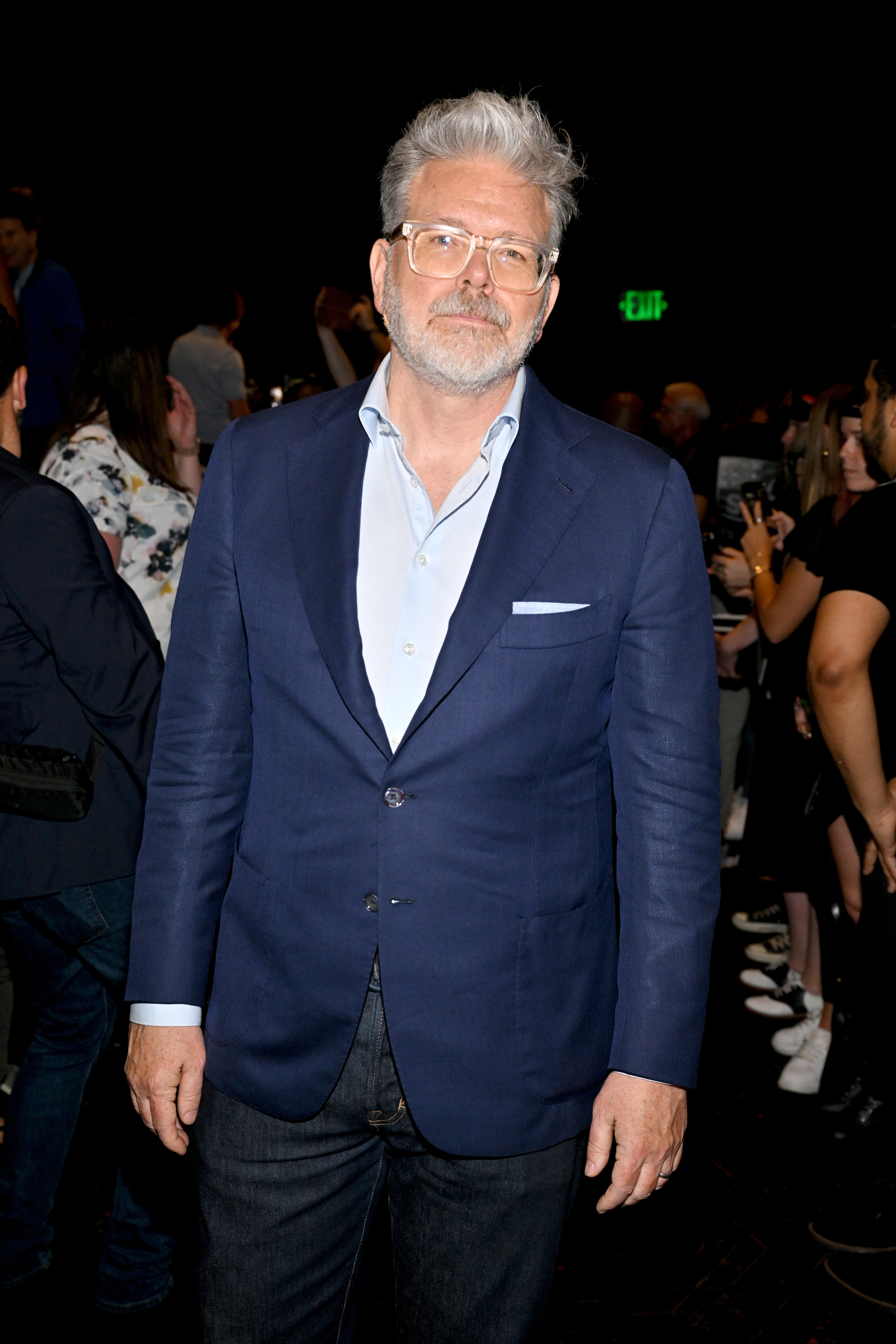 Christopher McQuarrie with gray hair and beard wears a blue blazer and light blue shirt at a public event. Other attendees are in the background