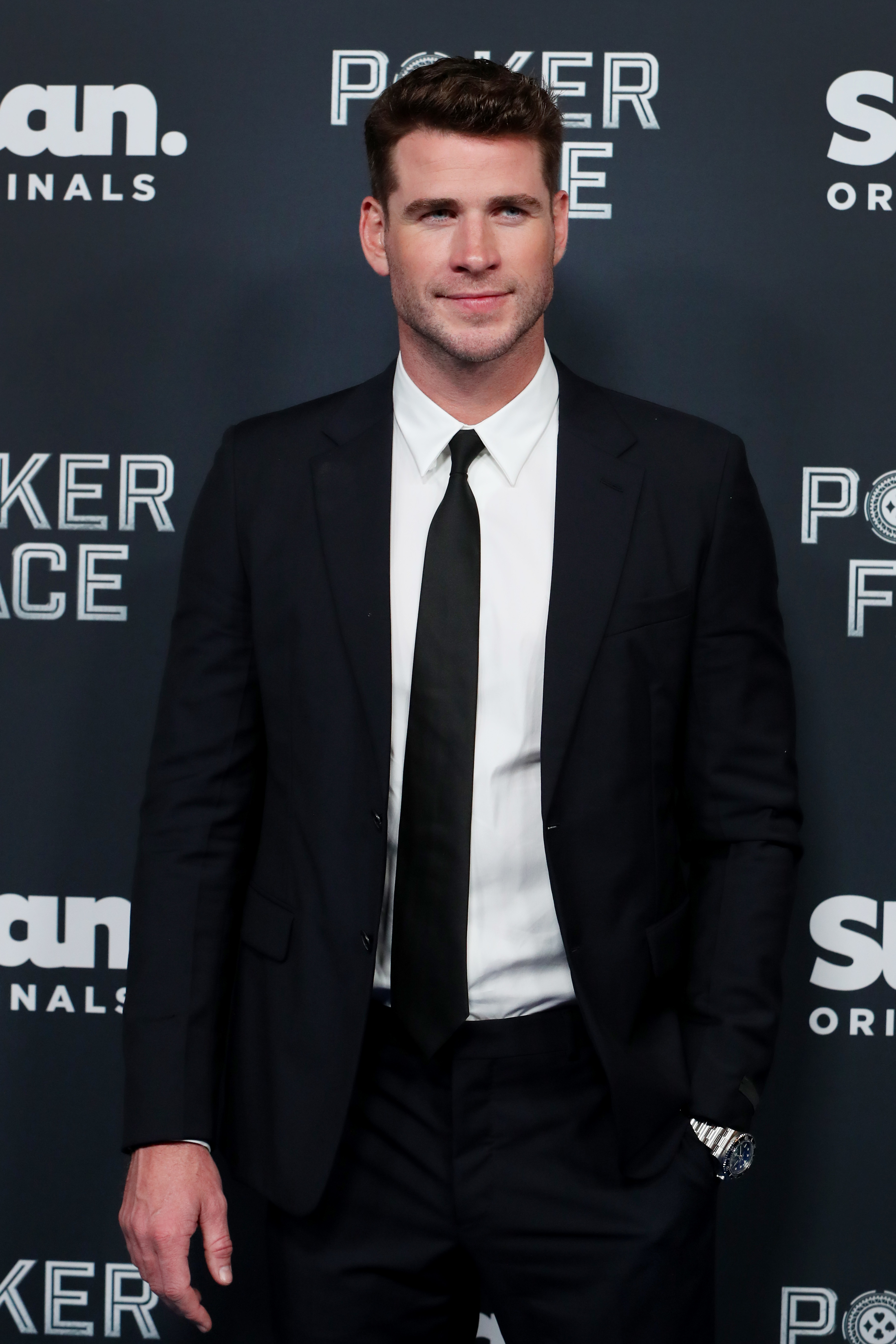 Liam Hemsworth at a &quot;Poker Face&quot; event, dressed in a classic black suit with a white shirt and black tie, posing in front of the event&#x27;s step-and-repeat backdrop