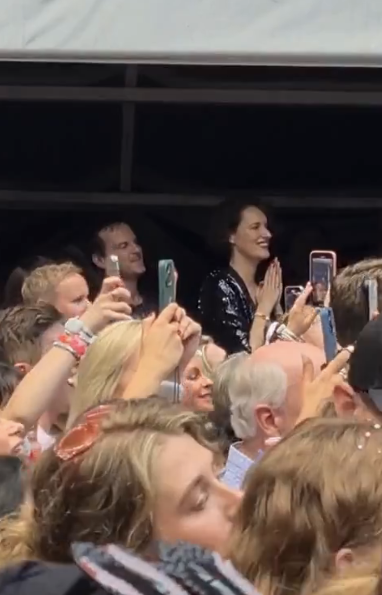 Phoebe Waller-Bridge stands in a crowd of people, smiling and clapping. Many attendees are holding up phones to take photos or videos
