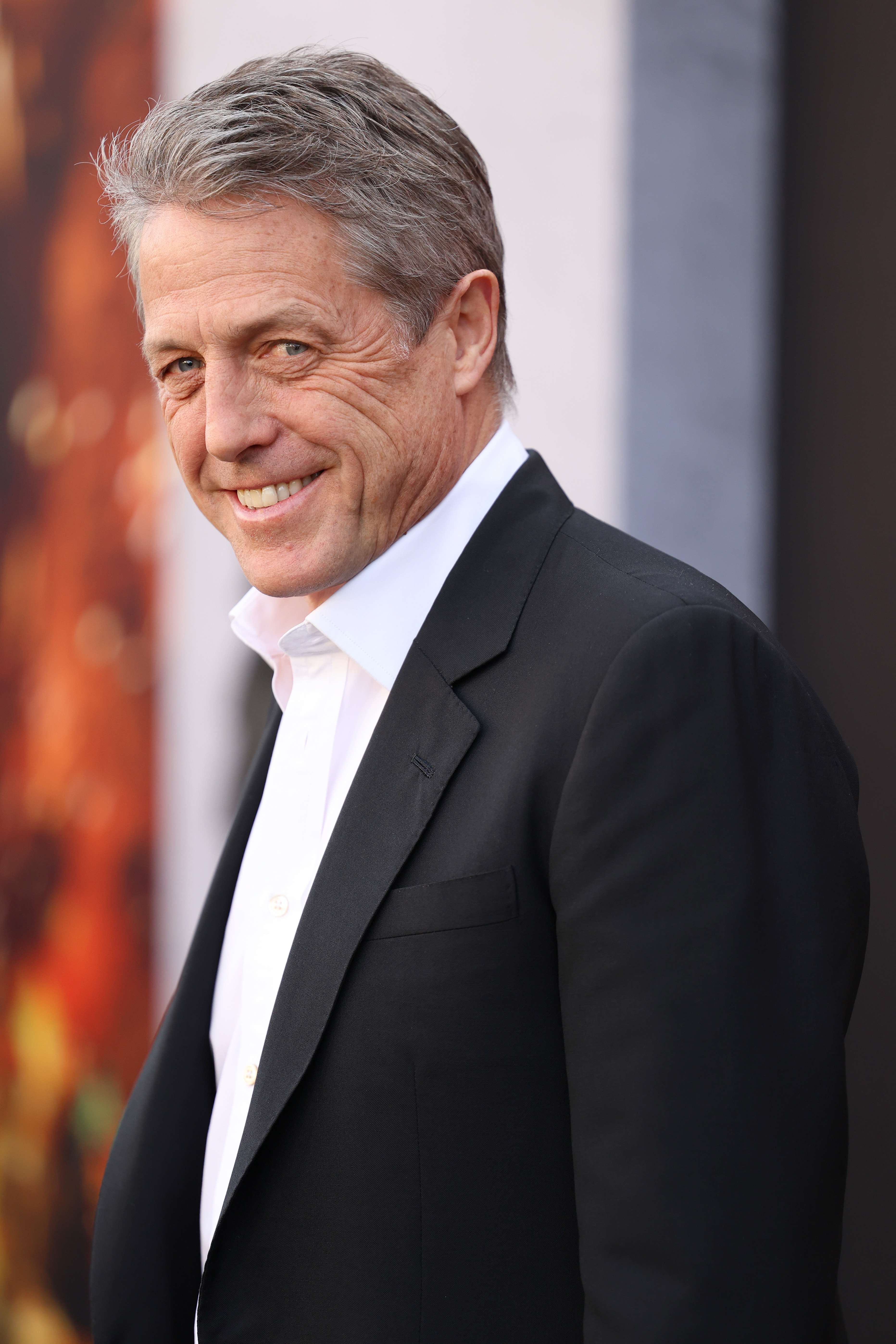 Hugh Grant smiles while wearing a black suit jacket over a white shirt
