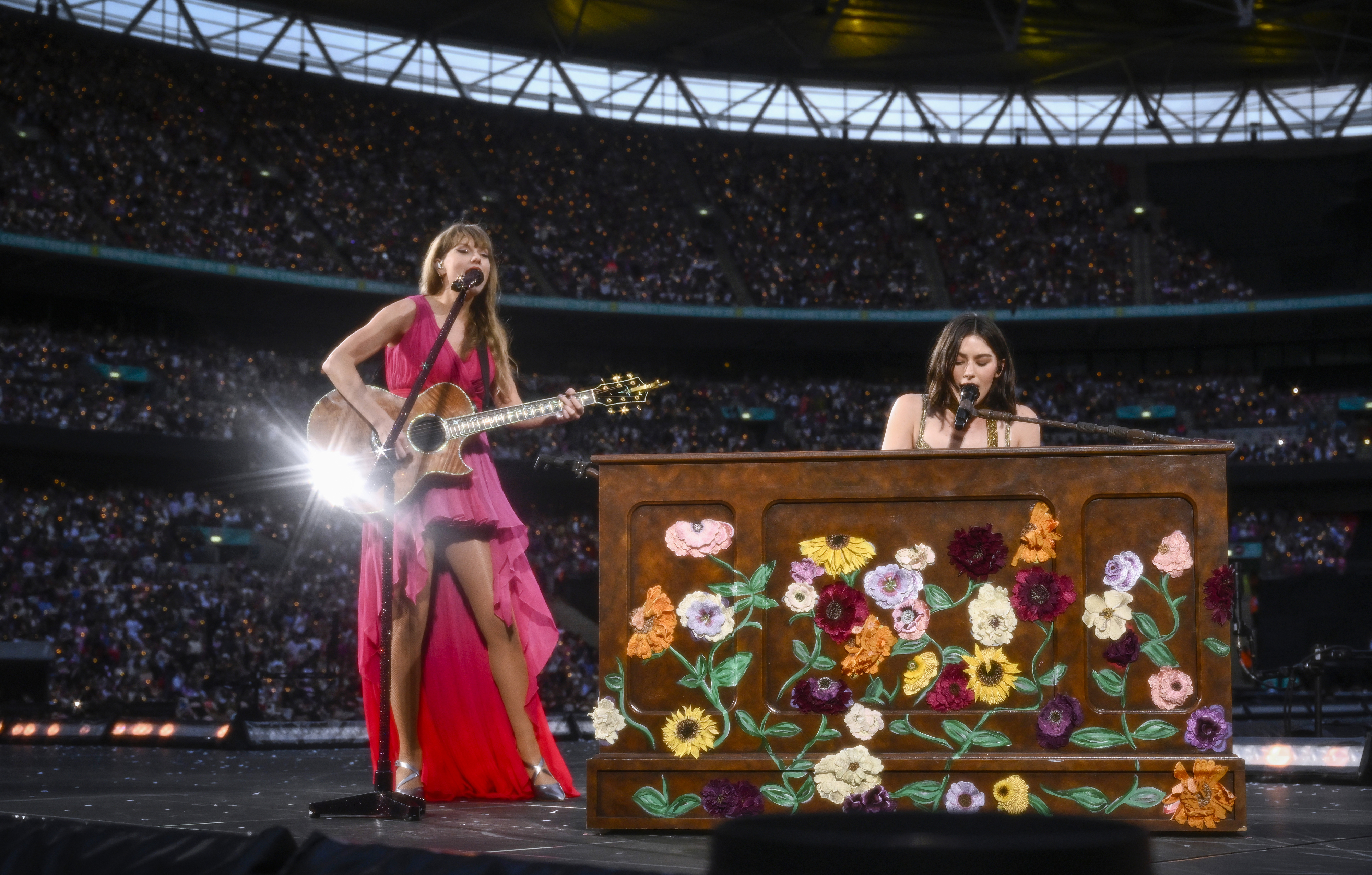 Taylor Swift, in a flowing dress with a high slit, sings and plays guitar on stage with Gracie Abrams playing a decorated piano at a concert