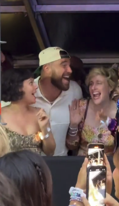 Three people, including Travis Kelce and Greta Gerwig, are smiling and laughing together at a crowded event with phones capturing the moment