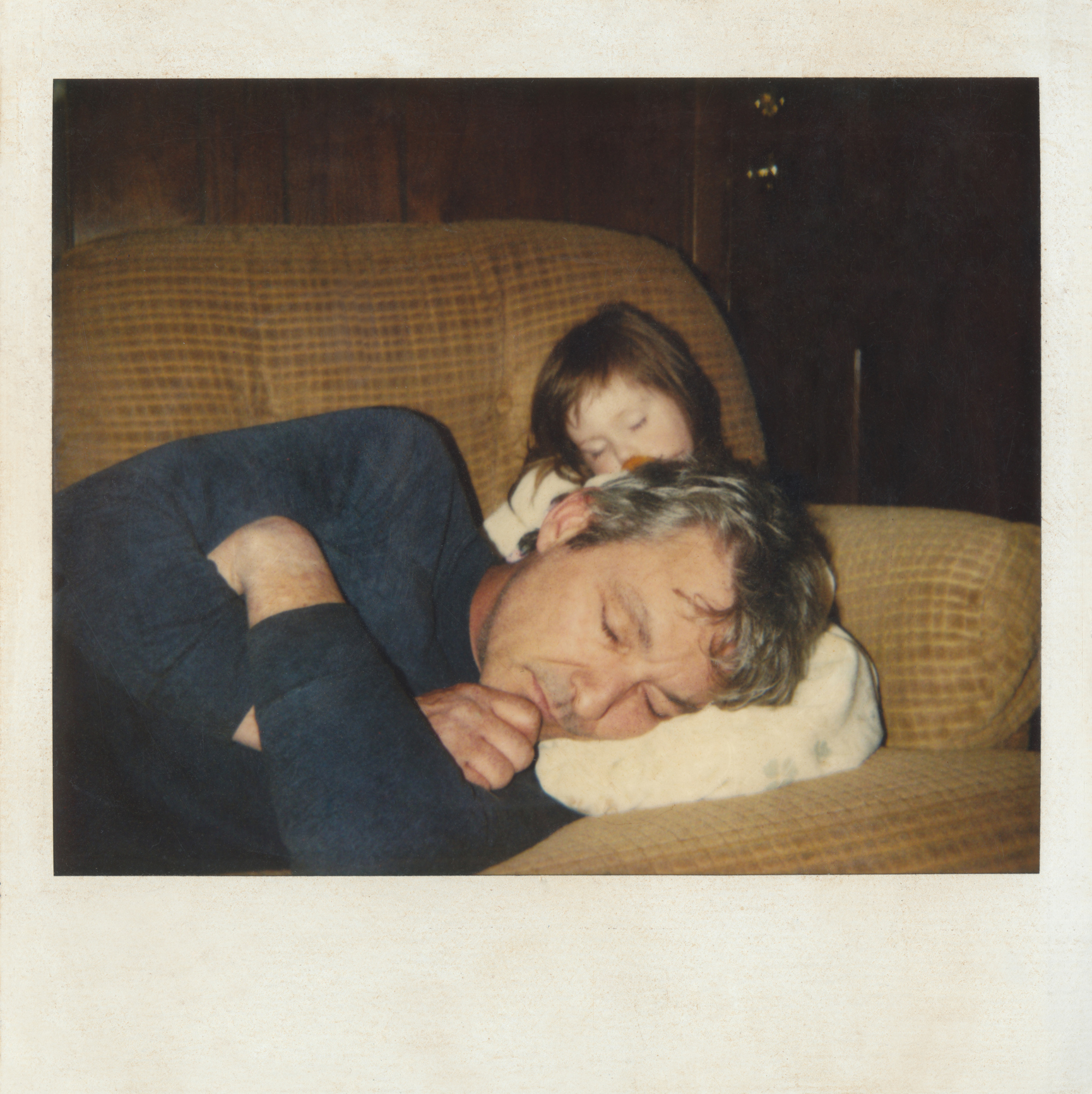 Man and young child sleeping on a couch, with the man's head resting on a cushion and the child resting on his shoulder