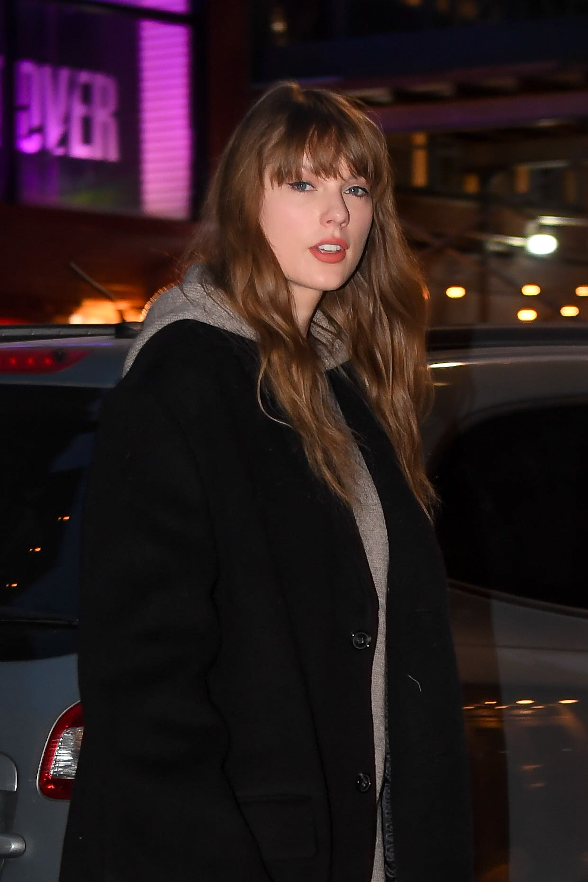 Taylor Swift walking outdoors at night in a casual hooded sweatshirt and a black coat