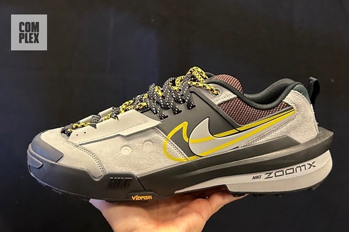 A person's hand holding a gray Nike sneaker with Vibram sole, yellow swoosh, and labeled "NIKE ZOOMX." The Complex logo is in the upper-left corner