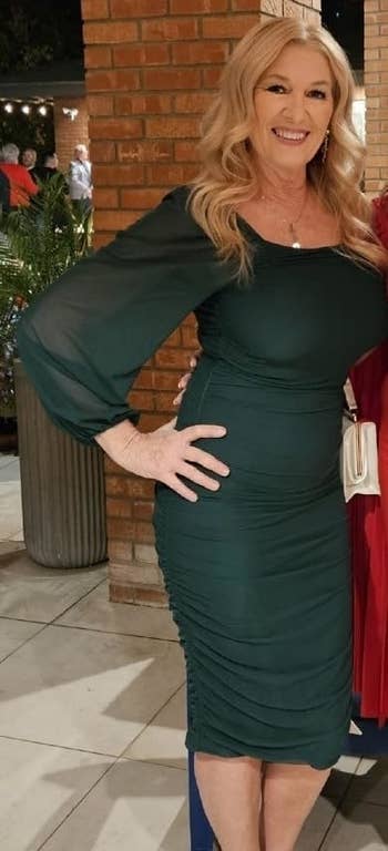 A smiling reviewer in a fitted, long-sleeve dress with ruched detailing poses with one hand on her hip in front of a brick wall and potted plants