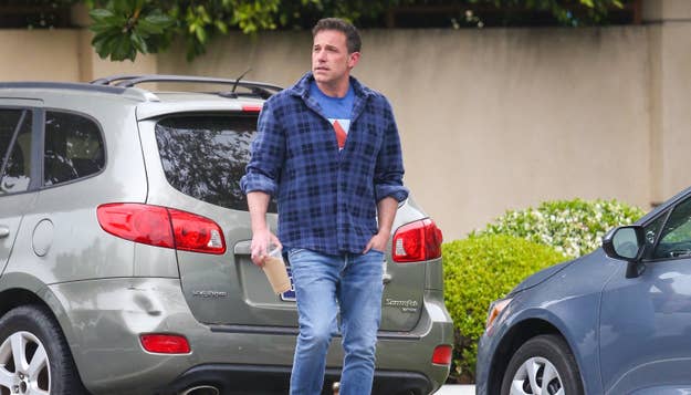 A man, not identified here, wearing a blue plaid shirt, jeans, and holding a coffee cup, walks near parked cars in a suburban area