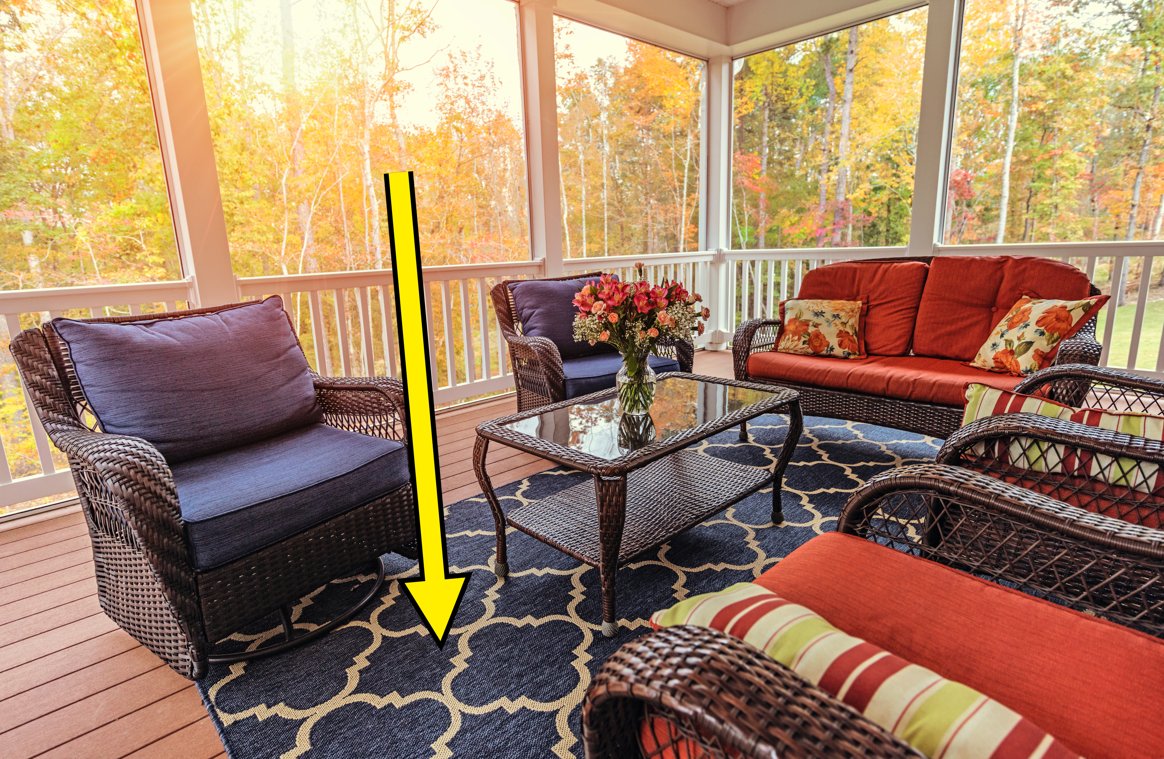 A cozy screened-in porch features wicker furniture with cushions and a glass-topped coffee table. Colorful throw pillows and a floral arrangement on the table add a decorative touch