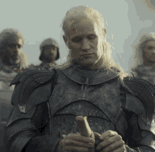Matt Smith, en su papel de un personaje medieval, viste armadura completa mientras observa una flecha en sus manos, rodeado de otros personajes en armadura