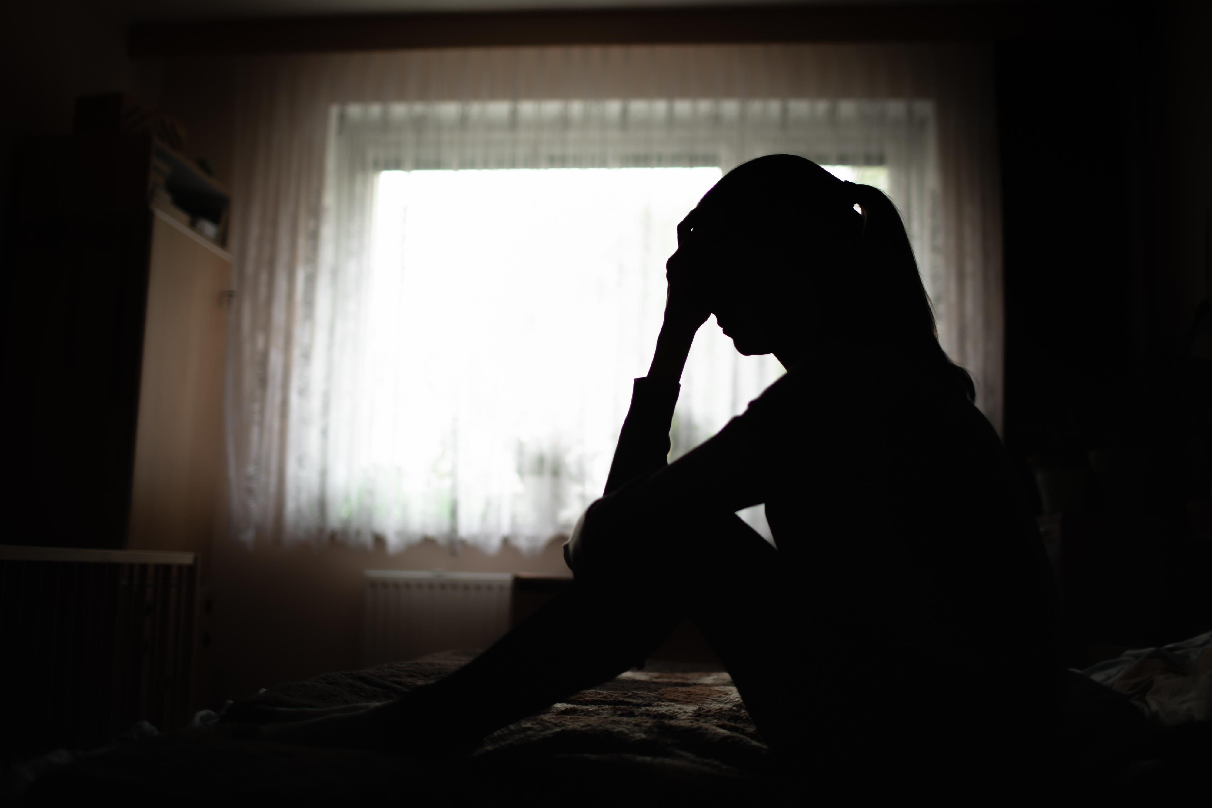 Silhouette of a person sitting on a bed with their head in hand in front of a window with sheer curtains