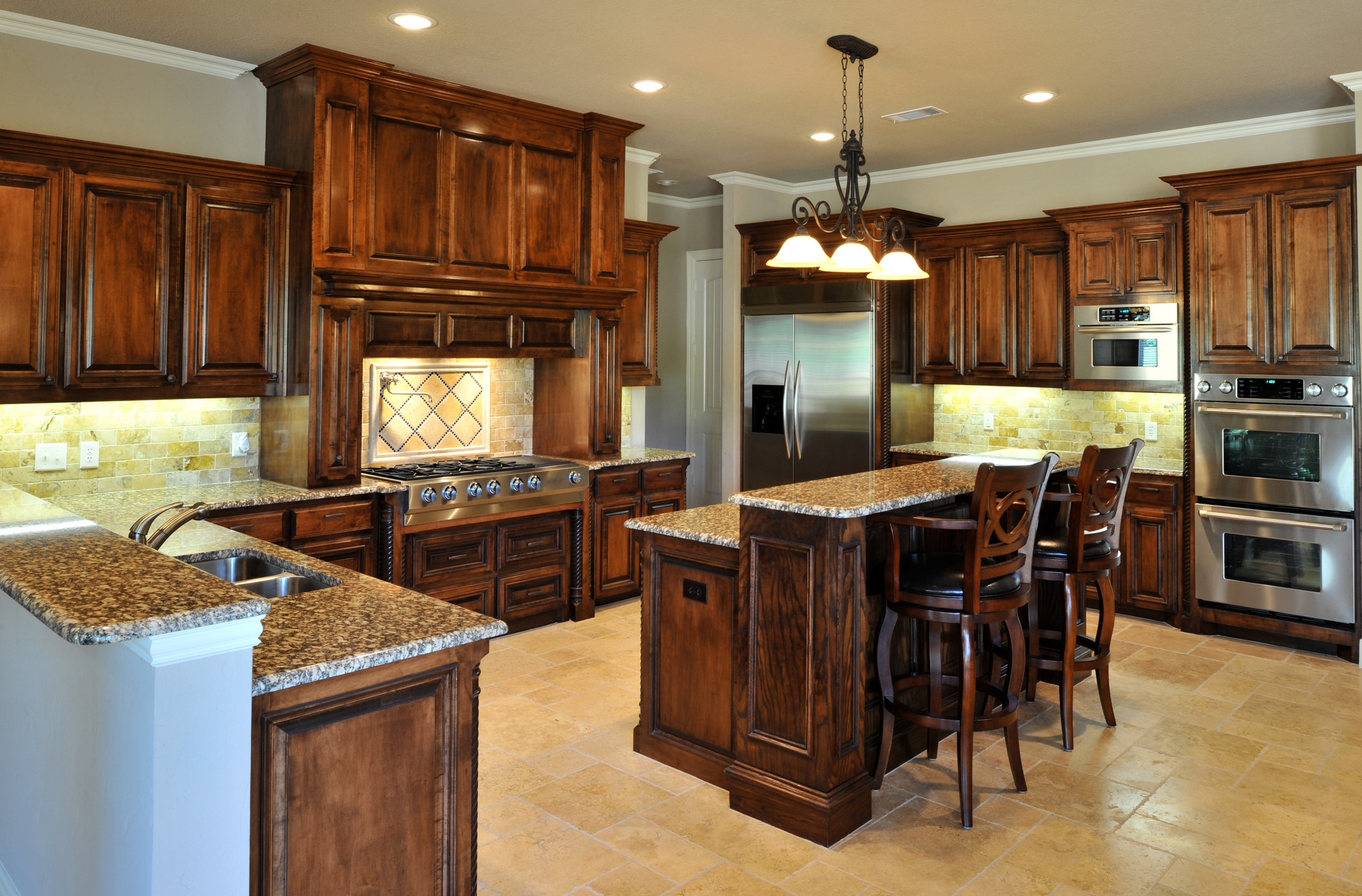 Spacious kitchen with wooden cabinets, granite countertops, stainless steel appliances, and an island with bar stools. Chandeliers hang from the ceiling