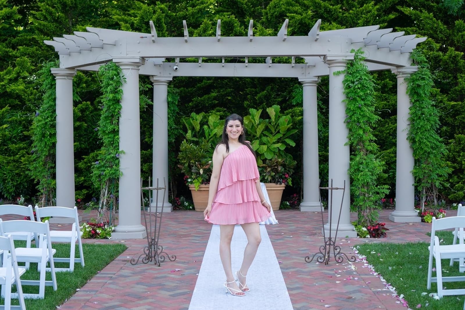 A reviewer stands under a pergola on a white aisle at an outdoor venue wearing a short, layered, sleeveless dress and white shoes