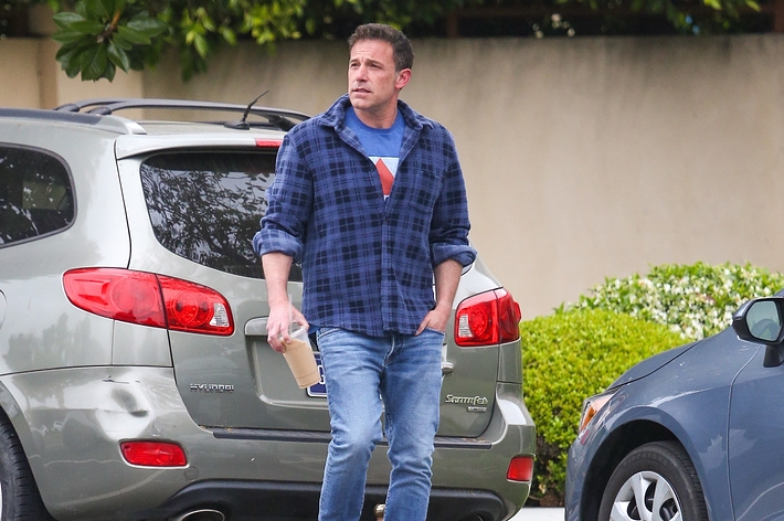 A man, not identified here, wearing a blue plaid shirt, jeans, and holding a coffee cup, walks near parked cars in a suburban area