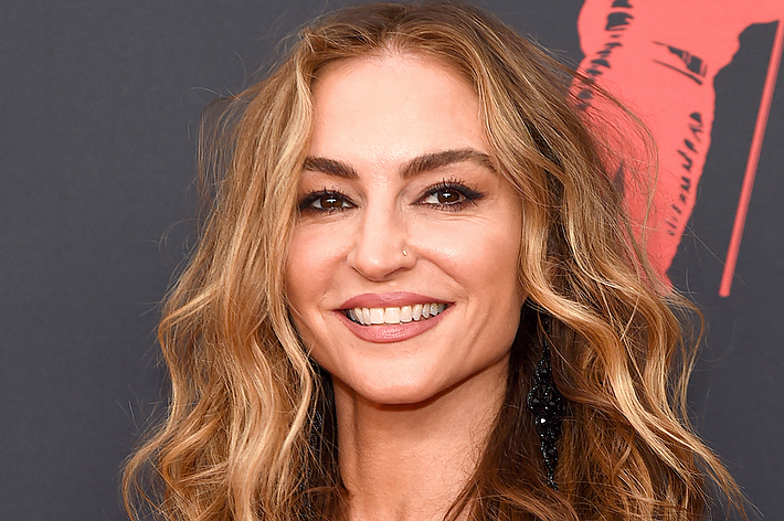 A smiling woman with wavy hair posing at a red carpet event, wearing a nose ring and black, sparkly earrings. Name unknown