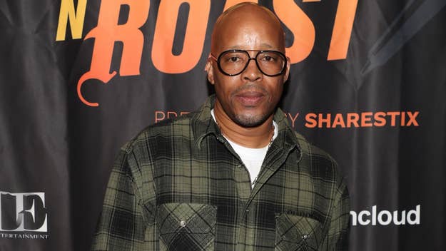 Portrait of Warren G on a music event red carpet wearing a plaid shirt and glasses against a backdrop with logos