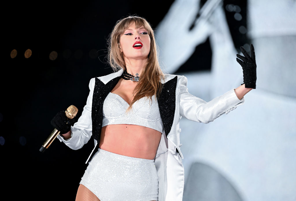 Taylor Swift performing on stage in a crop top and shorts outfit, holding a microphone and gesturing to the audience