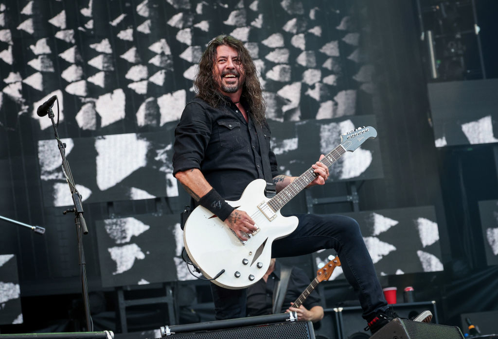 Dave Grohl performs energetically on stage with his guitar, wearing a button up shirt and pants. He smiles broadly against a backdrop of abstract designs