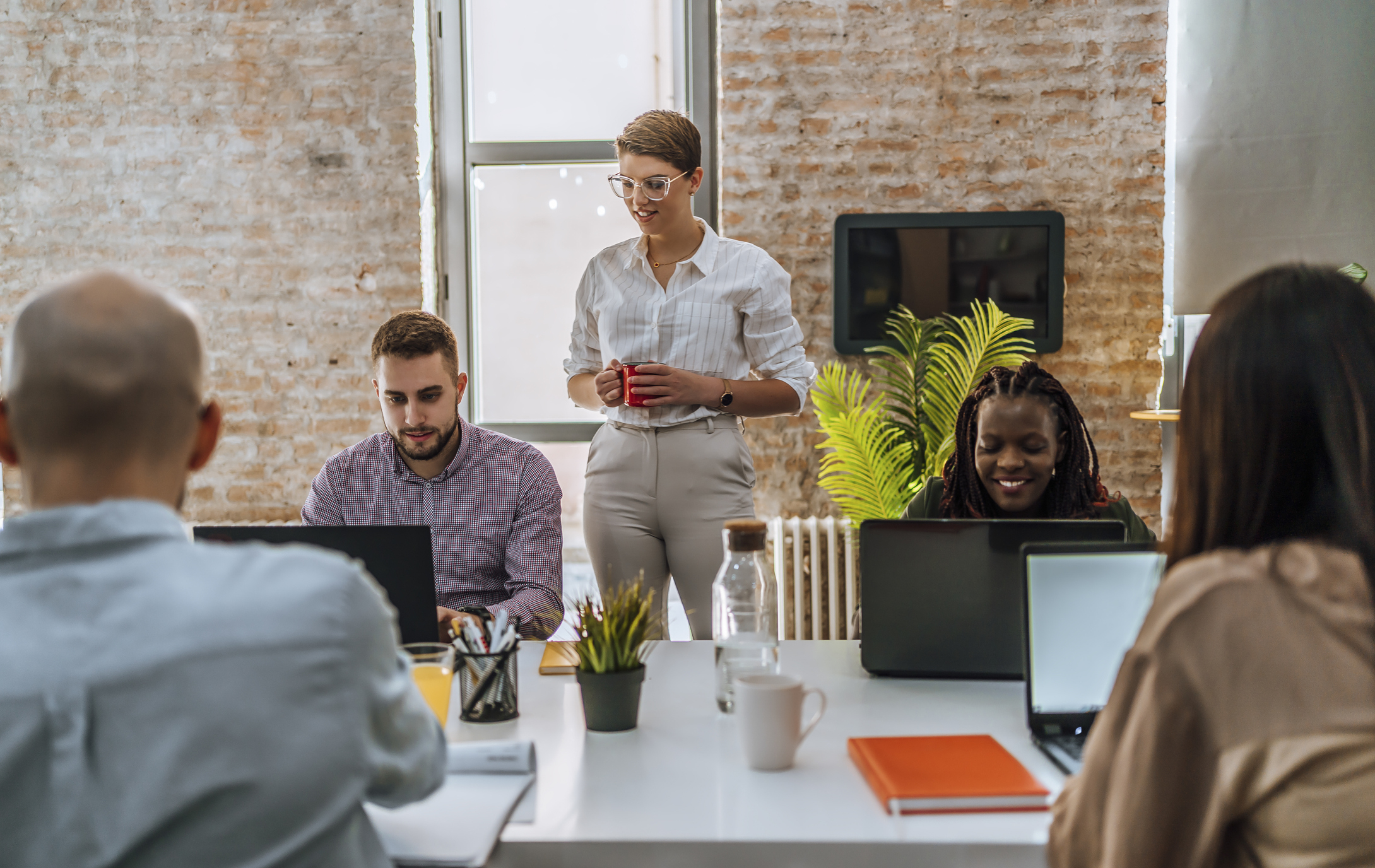 A group of five individuals is working together in an office setting. One person is standing with a mug, while the others are seated and using laptops