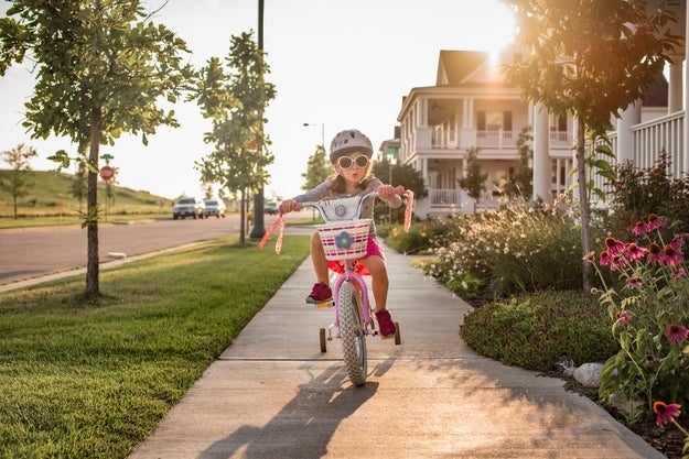 A young child rides a tricycle down a suburban sidewalk, wearing a helmet and sunglasses. Houses and gardens line the street with cars parked in the distance