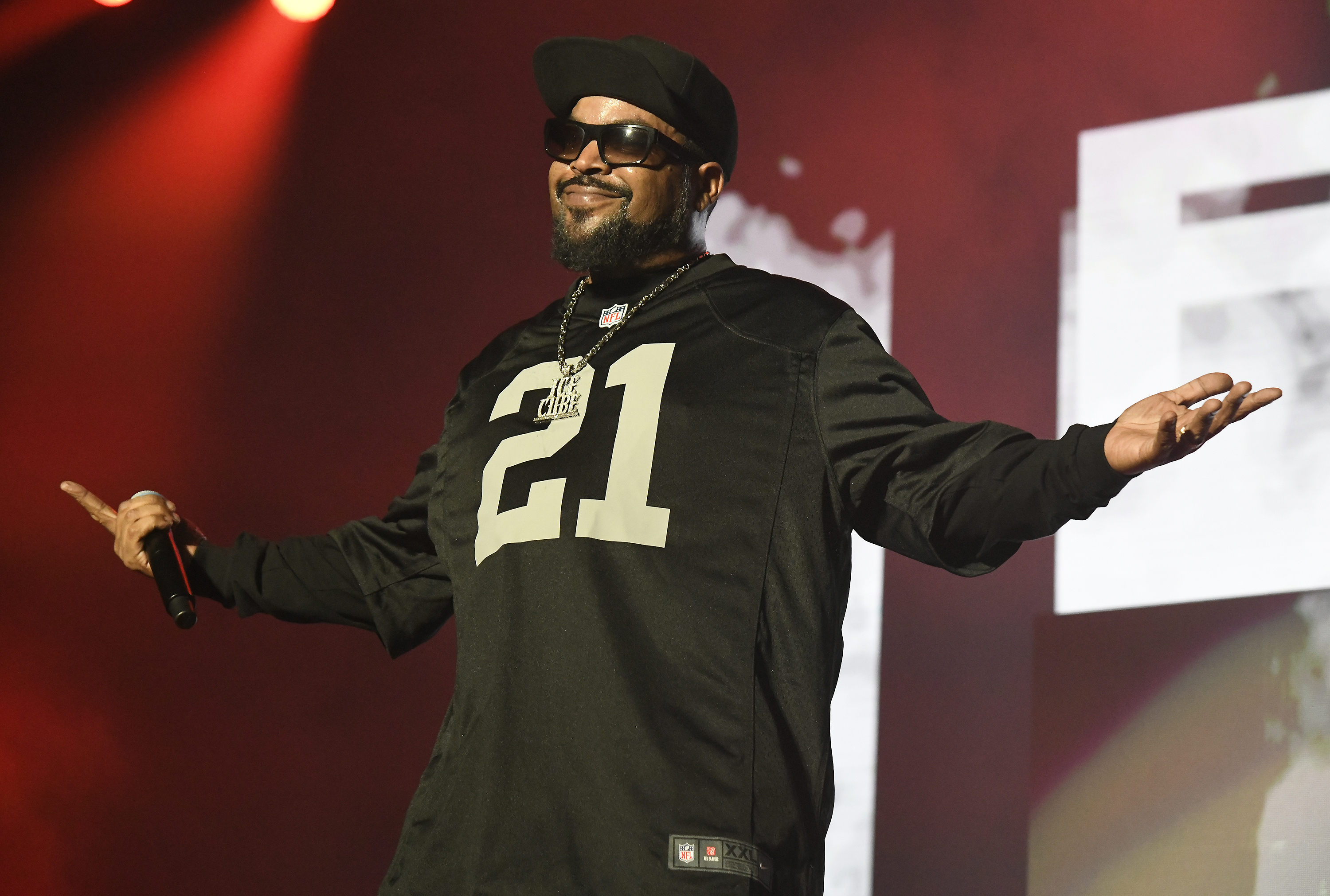 Ice Cube performing on stage, wearing a black sports jersey and dark sunglasses