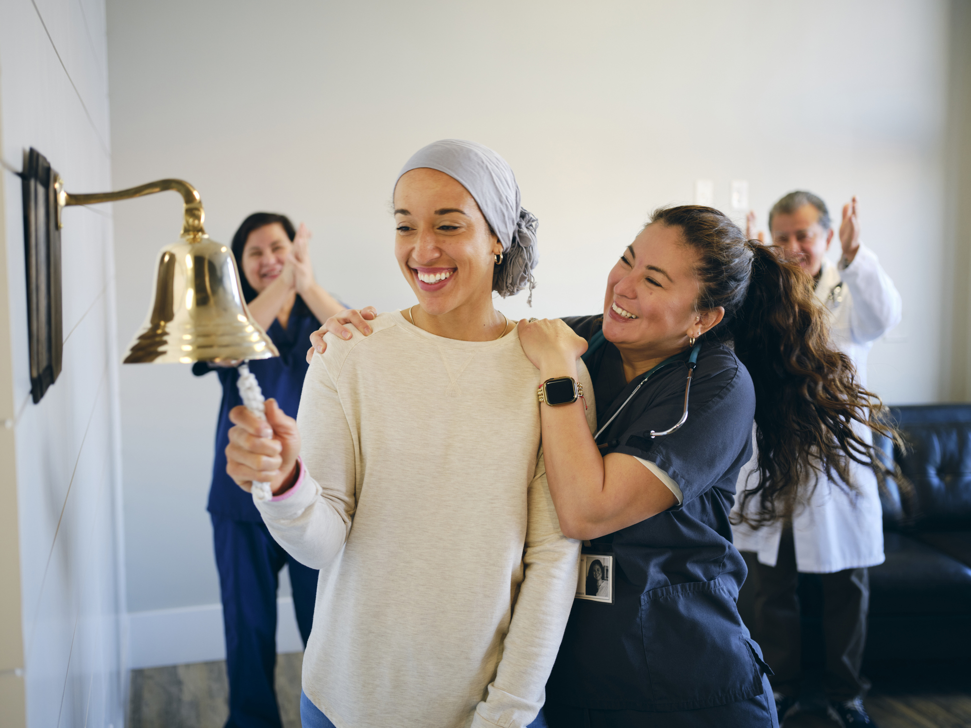 A woman wearing a headscarf smiles while ringing a bell. A nurse and two medical professionals in the background cheer and applaud her