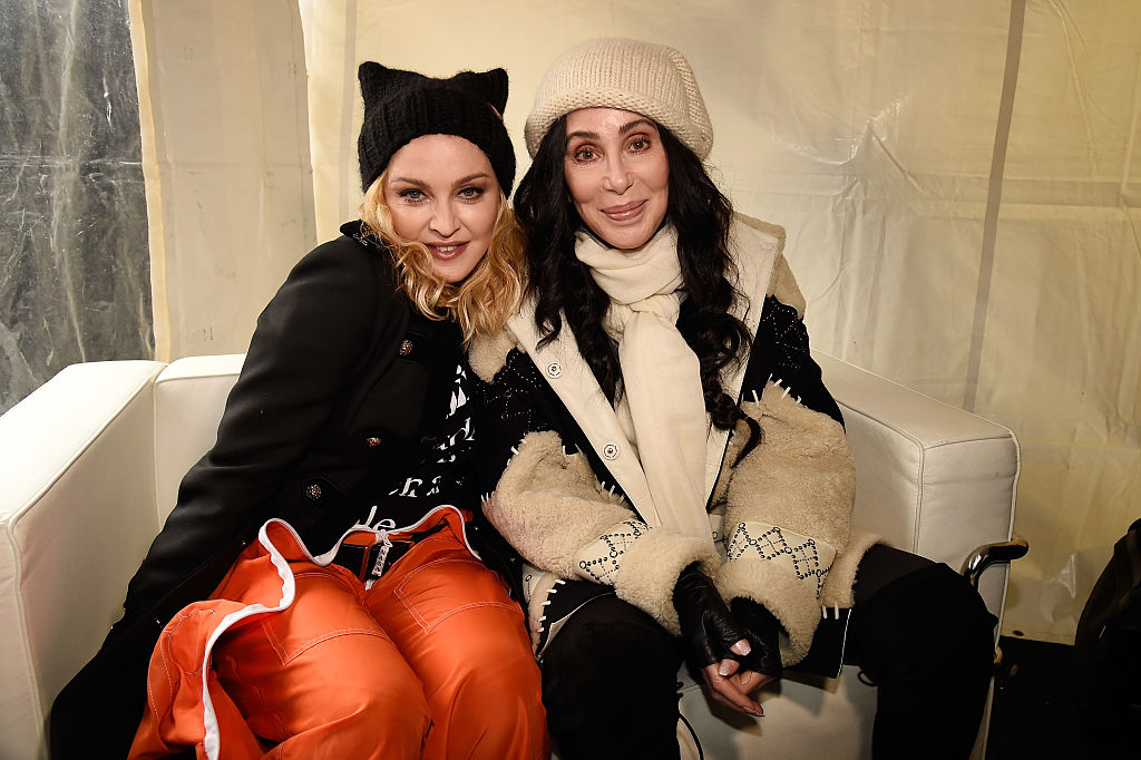 Madonna and Cher seated together, Madonna in a coat and hat with bright pants, Cher in a white knit hat and scarf with a coat