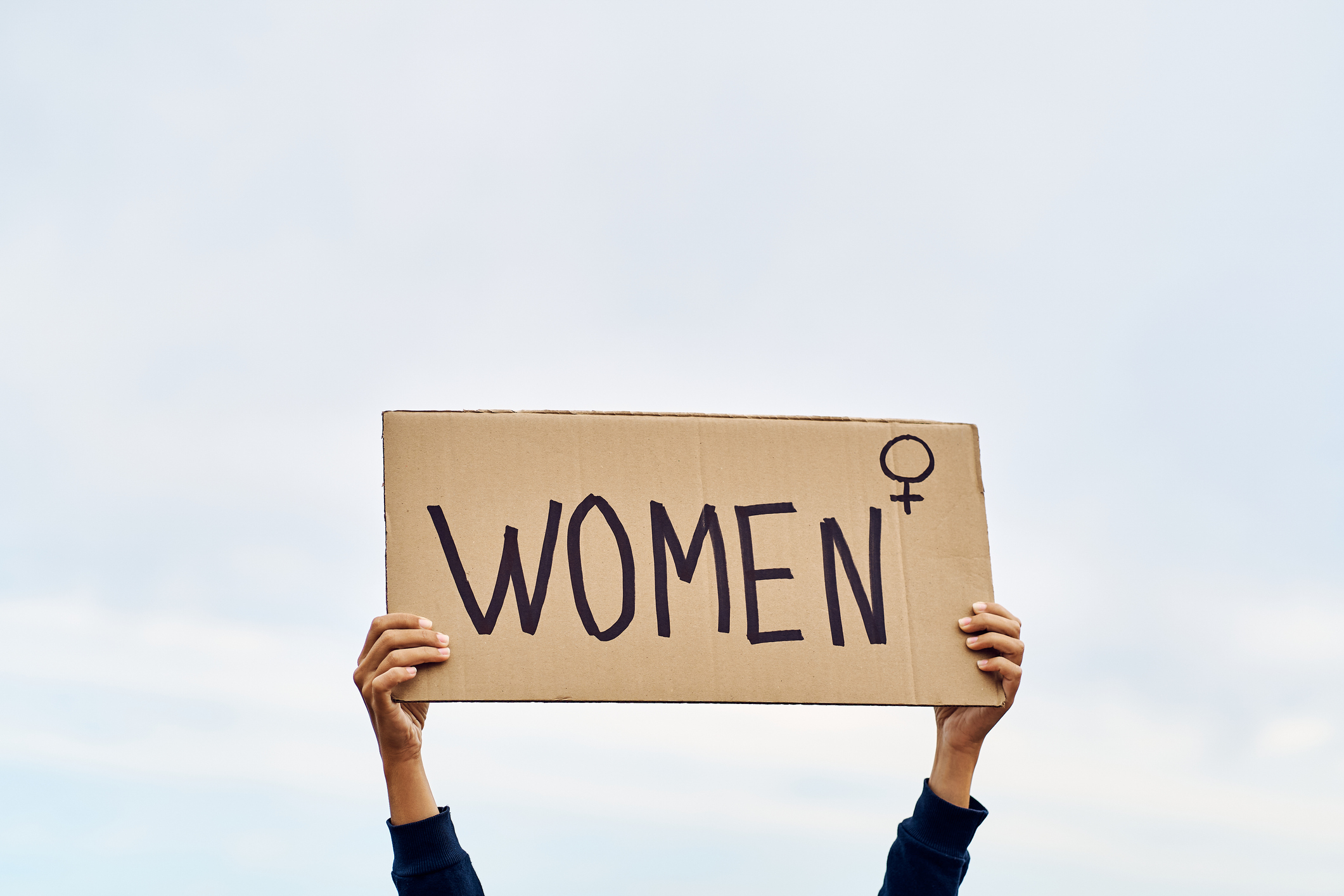 Two hands holding a cardboard sign that reads "WOMEN" with a female symbol next to it