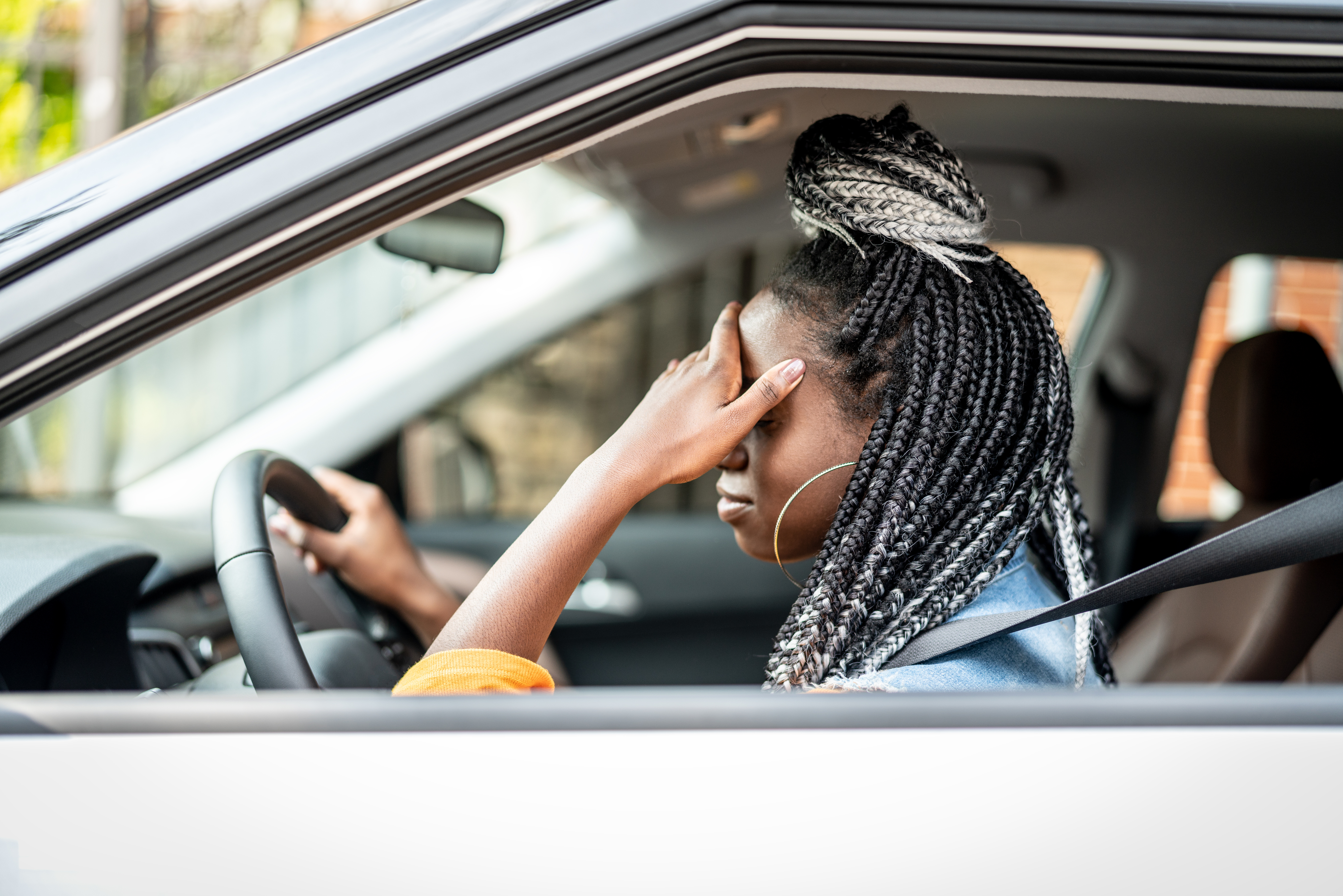 A person with braided hair is sitting in a car, holding the steering wheel with one hand while the other hand covers their face, appearing stressed