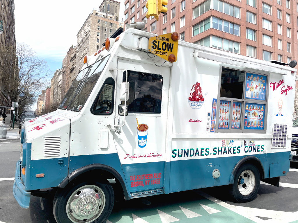An ice cream truck parked on a city street with a sign reading "Children Slow Crossing" on top. The truck offers sundaes, shakes, and cones