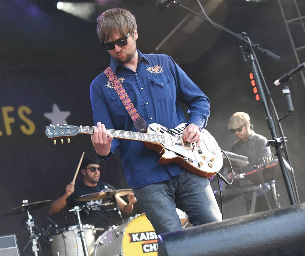Ricky Wilson of Kaiser Chiefs, in a patterned shirt and sunglasses, playing guitar on stage with bandmates on drums and keyboard