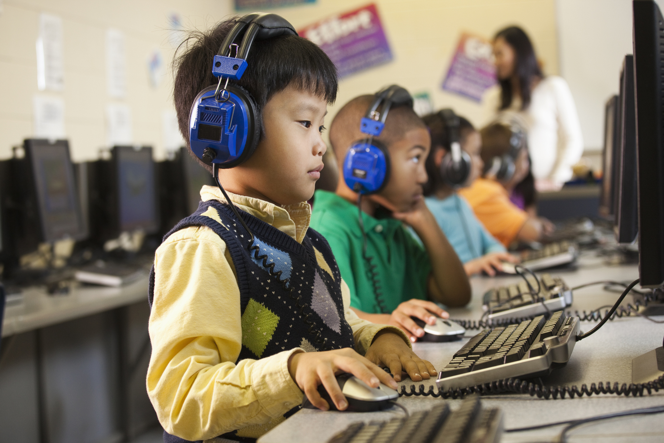 Children wearing headphones and using computers in a classroom setting. In the background, a teacher assists another student