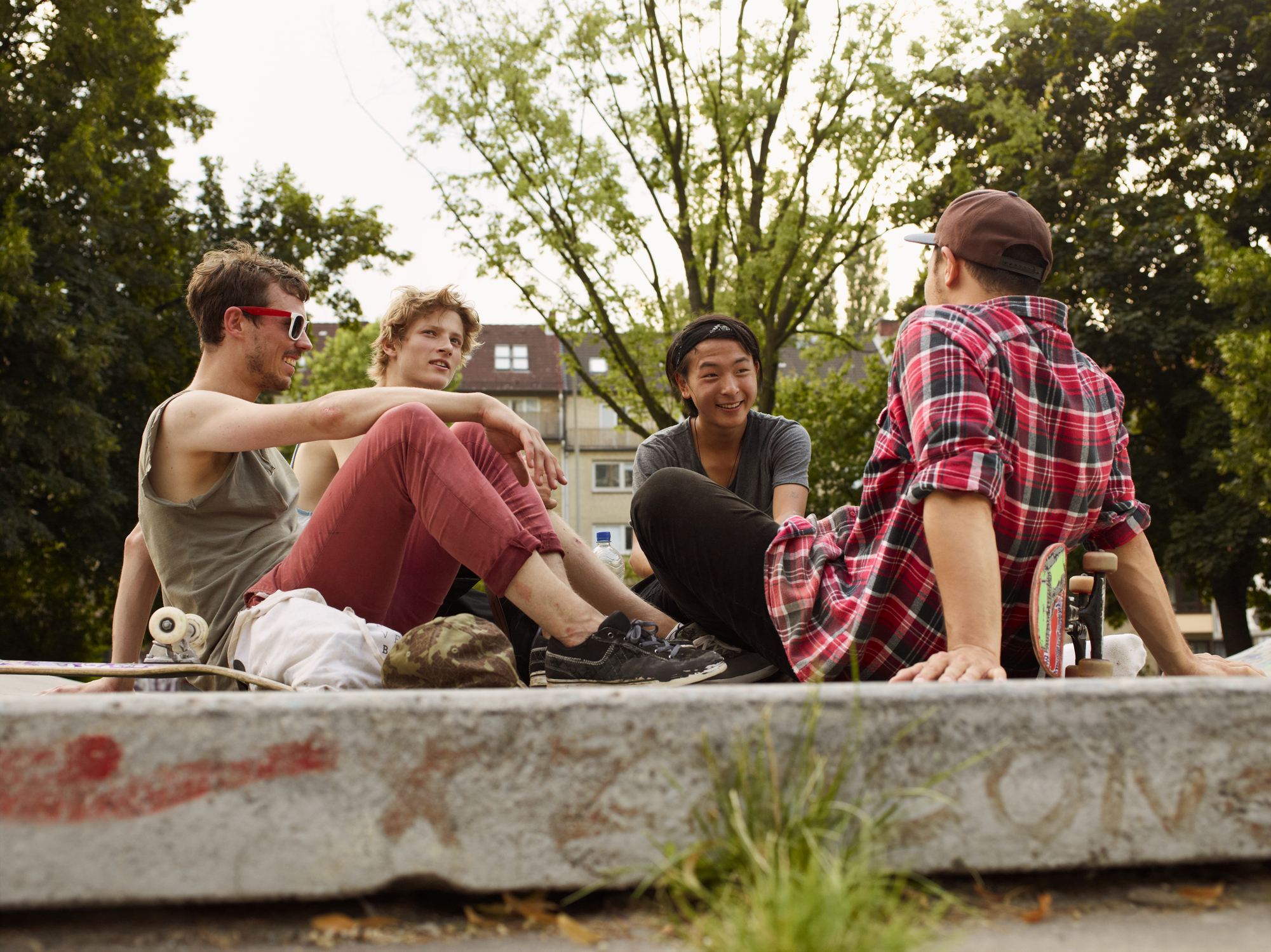 Four people sitting on a concrete ledge outdoors, chatting and relaxing. One is wearing a red plaid shirt and hat, others are in casual attire with skateboards nearby