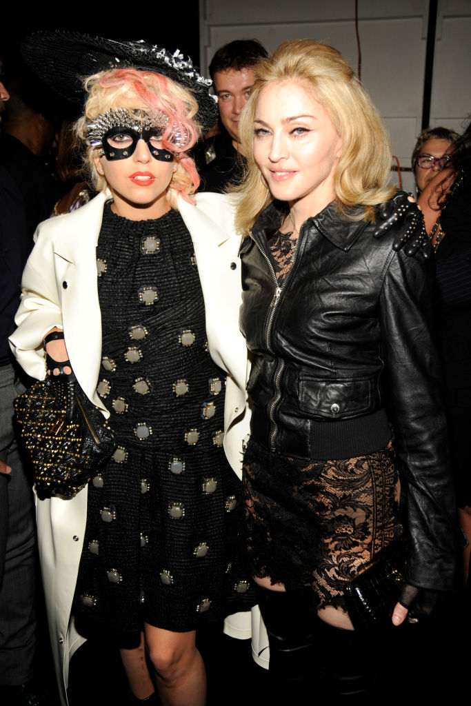 Lady Gaga and Madonna pose together. Lady Gaga wears an embellished dress with a trench coat and unique headpiece. Madonna wears a leather jacket and lace dress