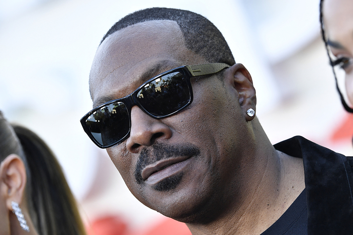 Eddie Murphy wearing sunglasses and a black jacket, posing on a red carpet